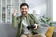 Joyful mature male celebrates a victory in a video game while sitting on a cozy sofa in a modern living room. Captures feelings of excitement and success.