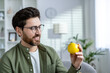 A cheerful bearded man with glasses smiles while holding a bright yellow apple in a modern living room setting, symbolizing healthy lifestyle choices.