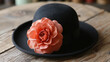A black fedora-style hat adorned with a large coral-colored artificial flower, placed on a wooden surface.