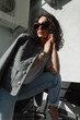 Stylish beautiful fashion curly haired woman model with trendy sunglasses in trendy casual clothes with blazer and t-shirt with jeans posing on the street at sunset