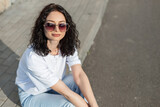 Fototapeta Panele - Summer stylish beautiful woman with curly hairstyle with heart-shaped sunglasses in a white T-shirt and jeans sits on the street and looks at the camera