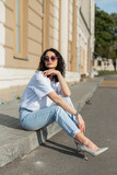 Fototapeta Panele - Stylish beautiful fashionable woman model with curly hairstyle with heart-shaped sunglasses in a fashion white T-shirt, jeans and shoes sits in the city