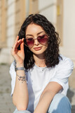 Fototapeta Panele - Fashion beautiful curly summer woman model in a white T-shirt puts on stylish heart sunglasses in the city in the sunlight