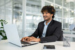 © Liubomir - A cheerful young male business professional engages with his laptop in a bright, contemporary office setting, exuding positivity and productivity.