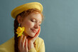 Fashionable happy smiling redhead freckled woman wearing yellow beret, silver earrings, holding daffodil flower, posing on blue background. Close up studio portrait. Copy, empty, blank space for text
