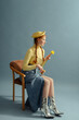 Fashionable redhead woman wearing trendy spring outfit with yellow beret, turtleneck, maxi blue denim skirt, silver ankle boots, posing on blue background. Studio full-length fashion portrait