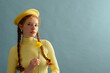 Fashionable confident redhead woman wearing yellow beret, silver earrings, ribbed turtleneck, holding daffodil flower, posing on blue background. Close up portrait. Copy, empty, blank space for text