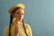 Fashionable confident redhead woman wearing yellow beret, silver earrings, ribbed turtleneck, holding daffodil flower, posing on blue background. Close up portrait. Copy, empty, blank space for text
