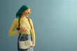 Fashionable confident redhead woman wearing green headscarf, yellow sunglasses, turtleneck, denim skirt, holding white leather purse, posing on blue background. Copy, empty, blank space for text