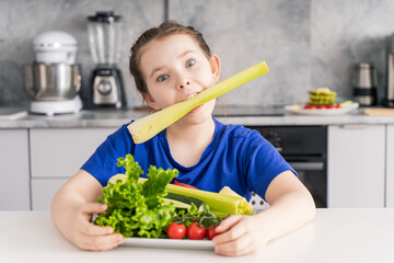 Cheerful little girl sitting with a plate of fresh vegetables and holding celery in her mouth. Cute girl having fun in the kitchen. Concept: natural vitamins in children's nutrition.