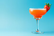 A vibrant orange colored margarita in an elegant glass, garnished with fresh strawberry and sugar rim on a blue background with copy space for text or product mockup.