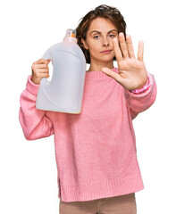 Poster - Young hispanic woman doing laundry holding detergent bottle with open hand doing stop sign with serious and confident expression, defense gesture