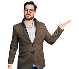 Wall Mural - Young hispanic man wearing business jacket and glasses smiling cheerful presenting and pointing with palm of hand looking at the camera.