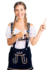 Beautiful caucasian woman with blonde hair wearing octoberfest traditional clothes smiling and looking at the camera pointing with two hands and fingers to the side.