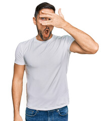 Sticker - Handsome man with beard wearing casual white t shirt peeking in shock covering face and eyes with hand, looking through fingers with embarrassed expression.