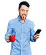 © Krakenimages.com - Hispanic man with beard using smartphone and drinking a cup of coffee sticking tongue out happy with funny expression.