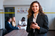 © artiemedvedev - Confident caucasian blonde businesswoman in smart casual attire uses smartphone in office, colleagues meet in background. Professional female checks email, manages tasks, smiles at camera.