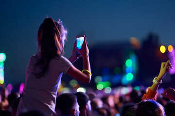 Wall Mural - Girl films music concert on smartphone amid lively festival crowd at twilight. Audience enjoys live performance, memories, vibrant stage lights beam. Fans wristbands share experience social media.