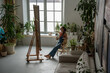 Satisfied artistic woman sitting relaxed on chair at easel holding brushes and palette with paints examining huge canvas with drawing. Female painter resting in middle of workflow, taking short break.