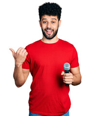 Wall Mural - Young arab man with beard singing song using microphone pointing thumb up to the side smiling happy with open mouth