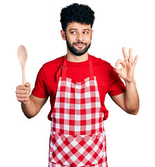 Sticker - Young arab man with beard wearing baker uniform holding wooden spoon smiling looking to the side and staring away thinking.