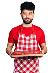 Poster - Young arab man with beard holding board with raw meat smiling looking to the side and staring away thinking.