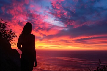 A young woman admiring a stunning sunset from a clifftop lookout, her silhouette outlined against the colorful evening sky