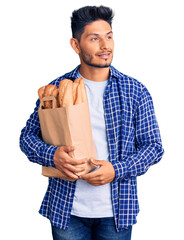 Canvas Print - Handsome latin american young man holding paper bag with bread smiling looking to the side and staring away thinking.