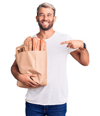 Sticker - Young handsome blond man holding paper bag with bread pointing finger to one self smiling happy and proud