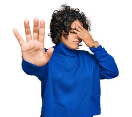 Canvas Print - Young hispanic woman with curly hair wearing turtleneck sweater covering eyes with hands and doing stop gesture with sad and fear expression. embarrassed and negative concept.