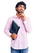 © Krakenimages.com - Handsome young man with curly hair and bear holding business folder serious face thinking about question with hand on chin, thoughtful about confusing idea