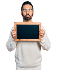 Canvas Print - Young hispanic man with beard holding blackboard making fish face with mouth and squinting eyes, crazy and comical.