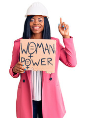 Poster - Young african american woman wearing architect helmet holding woman power banner surprised with an idea or question pointing finger with happy face, number one