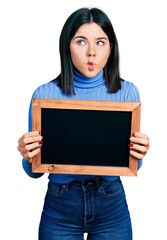 Poster - Young brunette woman with blue eyes holding blackboard making fish face with mouth and squinting eyes, crazy and comical.