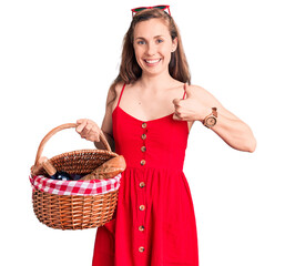 Sticker - Young beautiful blonde woman holding picnic basket smiling happy and positive, thumb up doing excellent and approval sign