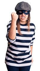 Canvas Print - Young beautiful brunette woman wearing burglar mask angry and mad raising fist frustrated and furious while shouting with anger. rage and aggressive concept.