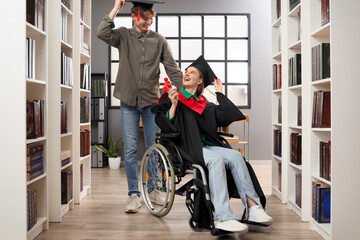 Wall Mural - Female graduate in wheelchair with diploma and her classmate at library