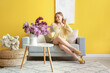 © Pixel-Shot - Young woman sitting on sofa with bouquets of lilacs at home