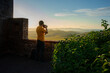 A photographer in a yellow shirt captures the sunset from an ancient wall, with a picturesque landscape in the background