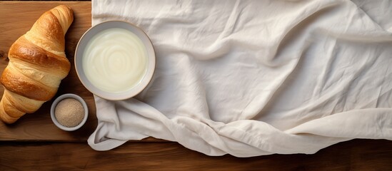 Poster - On a wooden table there is a top view of a fresh croissant placed on white fabric Additionally there is an egg in a paper panel and a rolling pin with flour in a bowl nearby creating a lovely copy sp