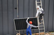 Men builders installing solar panel system on roof of house. Electricians in helmets lifting up photovoltaic solar module with help of ropes outdoors. Concept of alternative and renewable energy.