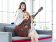 © Bangkok Click Studio - Two asian couple beautiful woman playing acoustic guitar, sing folk song together sitting on sofa at home, happy moments relaxing and enjoying lovers music and art therapy for people relax time.
