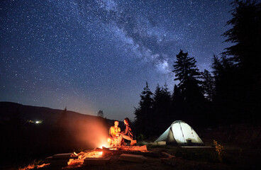 Wall Mural - Night-time camping under the twinkling stars. Man and woman, enthusiastic travelers, rejuvenate by campfire, near tent pitched amidst the tranquil woods, under the spellbinding night sky.