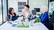 © makibestphoto - Trio of businesspeople, including a middle-aged Asian man and woman, strategize at their desk, prioritizing sustainability renewable energy adoption, waste reduction, and eco-friendly practices.