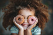 Portrait of a little smiling girl with curly hair and two appetizing donuts in her hands, closes her eyes with donuts. Happy National donut day concept