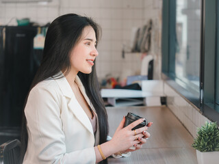 Young beautiful Asian businesswoman smiling and posing while sitting in a modern coffee shop. Young attractive woman sitting in an indoor cafe and looking happy at the camera.