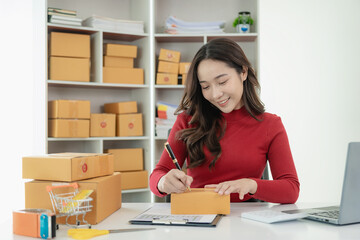 Wall Mural - Young Asian business woman smiles and uses laptop to check information on shipping box before sending it to customer. Small business entrepreneur, SME online marketing, delivery concept