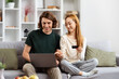 Young Couple Shopping Online In Living Room. A cheerful couple comfortably seated on a couch, browsing on a laptop, and holding a credit card, reflecting a happy online shopping experience.