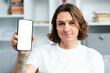 Smiling Young Man In White T-Shirt Showing Blank Smartphone Screen Indoors, Concept Of Technology, Communication, And Modern Lifestyle