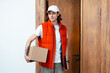 Friendly Delivery Man Holding Package By Wooden Door. Young Male Courier In Red Vest And White Cap Smiling, Ready For Efficient Service And Fast Shipping Concept.